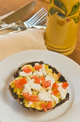top view, close distance of a portobello mushroom cap filled with scrambled eggs, tomato chunks, cheese and basil, on round, white plate, with olive oil ceramic bottle and yellow napkin
