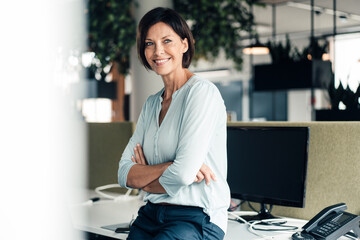Smiling female entrepreneur with arms crossed against computer in office