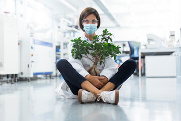 Mature female scientist with potted plant sitting in laboratory during COVID-19
