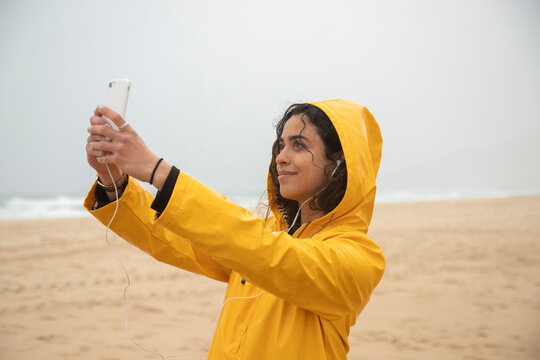 Smiling woman wearing yellow raincoat taking selfie through mobile phone while standing at beach