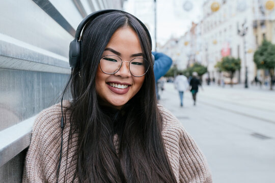 Smiling woman with headphones leaning on metal wall in city