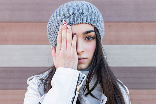 Teenage girl in knit hat covering her eyes with hands against wall