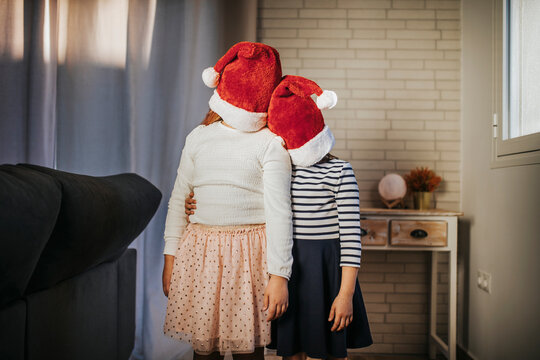Sisters covering their faces with Santa hat while standing in living room