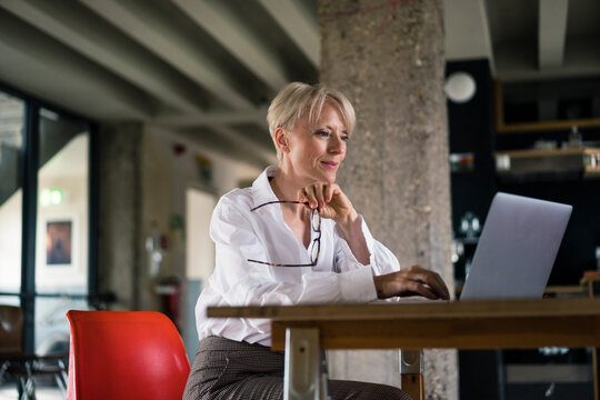 Smiling businesswoman holding eyeglasses while using laptop at desk by column at home office