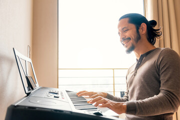 Young latin man learning to play piano at home in the living room with a window light