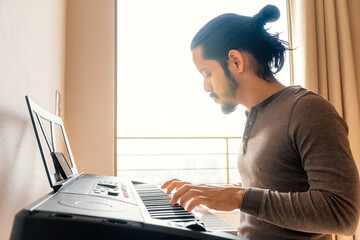 Young latin man learning to play guitar at home in the living room with a window light