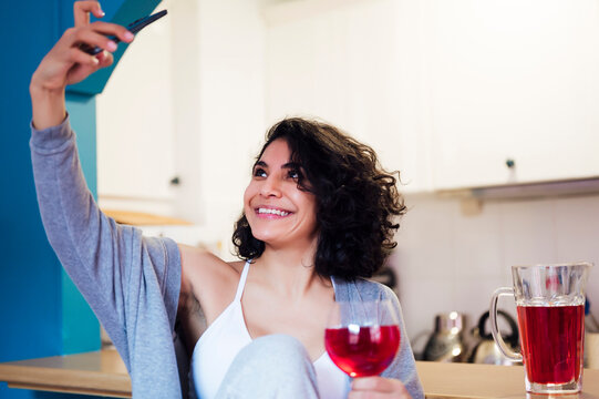 Smiling woman holding glass of juice while taking selfie through mobile phone at kitchen