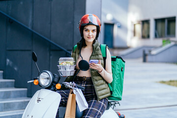 Delivery woman in red helmet with phone and a delivery motorcycle holding conteiners with lunch, outdoor. E-commerce concept, delivery service on mobile application.