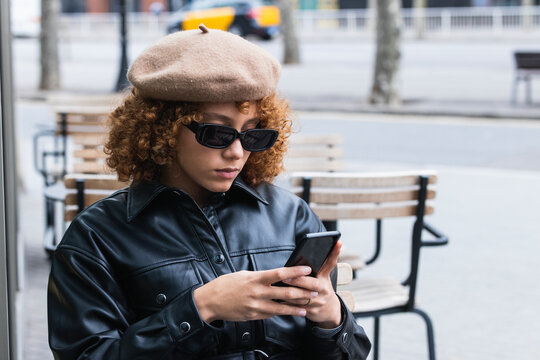 Fashionable teenage girl wearing beret and leather jacket using mobile phone at sidewalk cafe