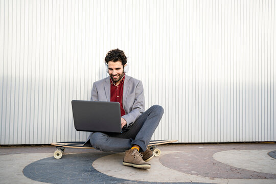 Smiling businessman with headphones using laptop while sitting on longboard in front of wall - Powered by Adobe