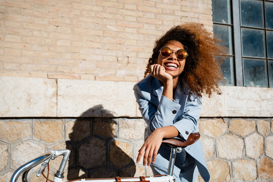 Cheerful young woman in sunglasses leaning on bicycle while standing against building during sunny day