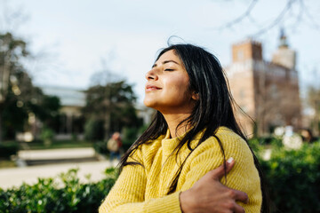 Smiling woman hugging self against sky in public park
