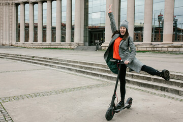 Cheerful woman with hand raised standing on electric push scooter near building