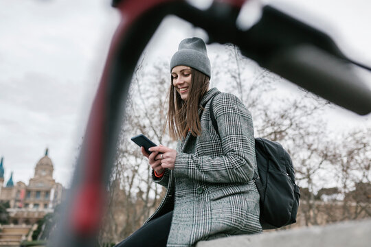Woman using smart phone while sitting on retaining wall in city