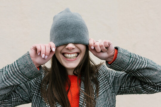 Smiling woman covering eyes through knit hat against wall