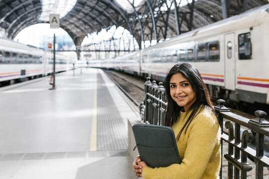 Smiling Mid Adult Woman With Laptop Bag At Railing On Platform