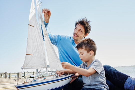 Father and son preparing toy sailboat together while sitting on pier