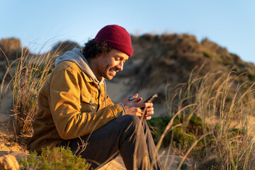 Smiling man wearing knit hat using smart phone while sitting by grass