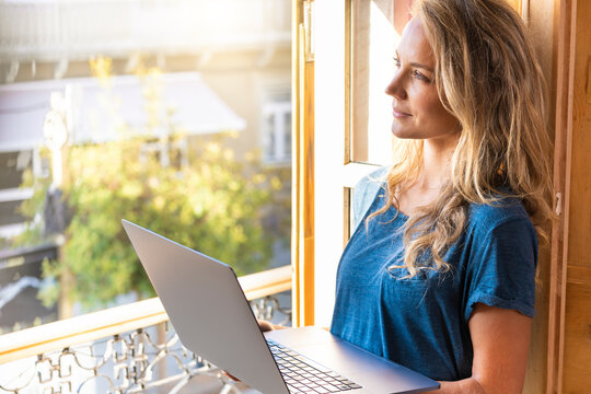 Woman with laptop contemplating while standing by window at home
