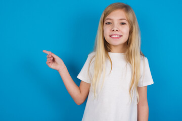 Positive beautiful Caucasian little girl wearing white T-shirt over blue background with satisfied expression indicates at upper right corner shows good offer suggests to click on link