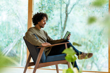 Young man using digital tablet while sitting on armchair by window at front yard