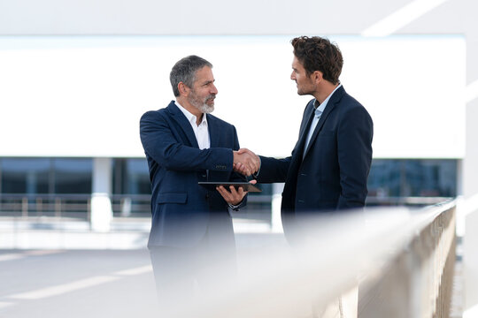 Mature entrepreneur holding digital tablet while doing handshake with colleague standing at office building terrace