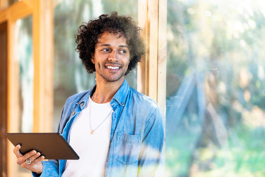 Smiling man with digital tablet looking away while leaning on window at front yard