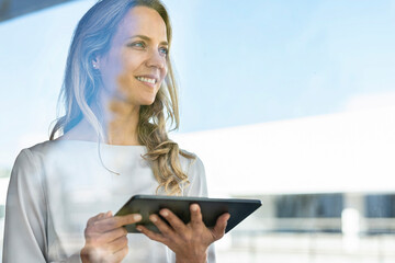 Smiling woman with digital tablet looking away while standing by window at office