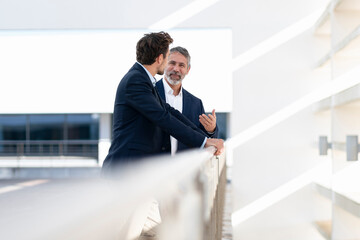 Mature businessman smiling while talking with colleague standing at office building terrace
