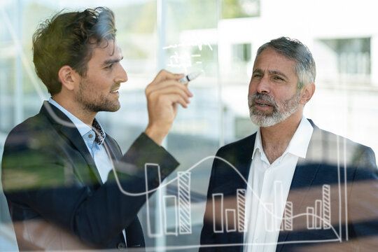 Male entrepreneur working with colleague on graph on glass at modern office