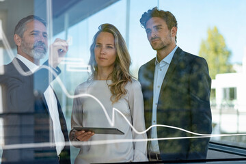 Male entrepreneur drawing preparing business plan on glass while standing with colleagues at office