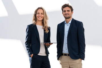 Confident businesswoman with digital tablet standing by colleague against white wall