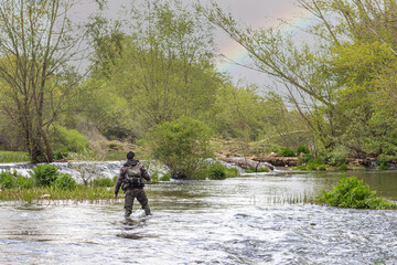 Boy fishing in the middle of a river with rocks, weeds and bushes with camouflage suit, long rubber boots, fishing rod and other tools for fishing with the rainbow in the sky - obrazy, fototapety, plakaty