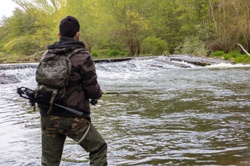 Naklejka premium Angler with camouflage clothing, backpack with fish dryer and rubber boots fishing with a rod in a river with a small waterfall in the background