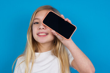 beautiful Caucasian little girl wearing white T-shirt over blue background holding modern smartphone covering one eye while smiling
