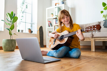Smiling redhead girl playing guitar while e-learning through laptop at home