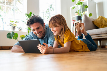 Cheerful father and daughter looking at digital tablet while lying down on floor in living room at home