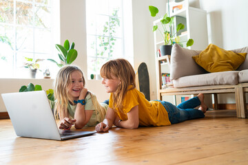 Smiling girls looking at each other while lying down on floor in front of laptop at home