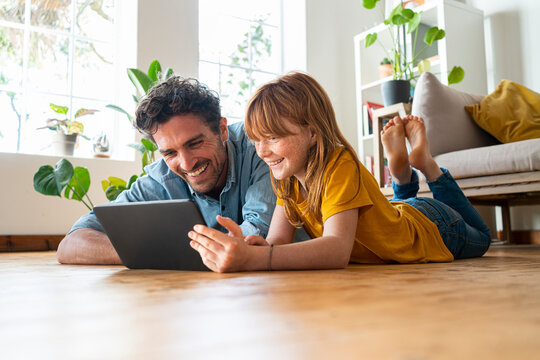 Cheerful father and daughter looking at digital tablet while lying down on floor in living room at home