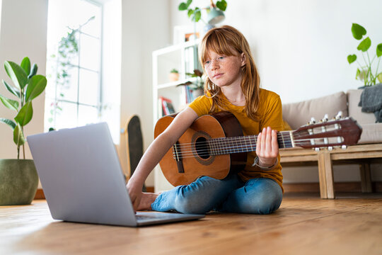 Redhead girl using laptop while learning guitar at home