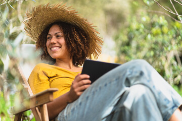 Smiling woman in straw hat holding digital tablet while sitting on chair in vegetable garden