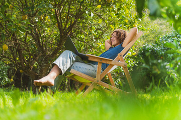 Smiling mid adult woman with hands behind back looking at laptop while sitting on chair in garden