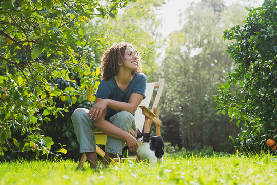 Smiling curly haired woman looking away while sitting on chair by cat in garden