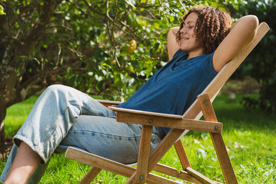 Smiling curly haired woman with eyes closed resting on deck chair in garden