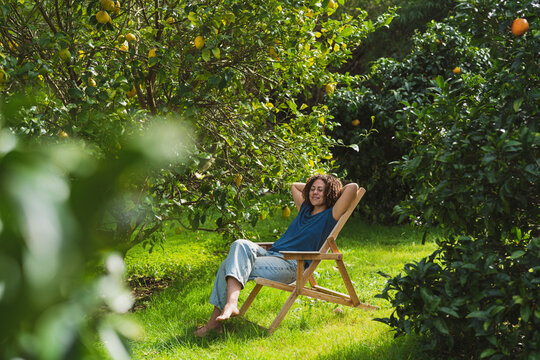 Woman with hands behind head relaxing on chair amidst trees in garden
