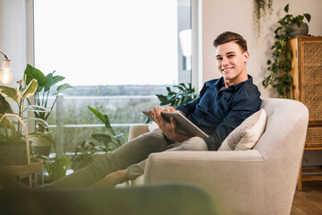Smiling young man with digital tablet sitting on sofa in living room