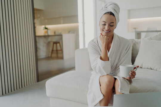 Happy woman in white bathrobe drinking tea and relaxing on sofa after taking shower or bath