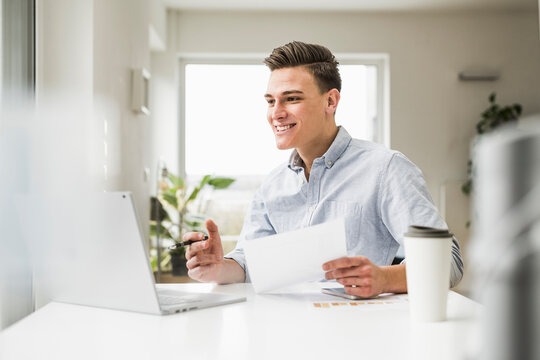 Happy businessman with document making video call through laptop sitting at desk - Powered by Adobe