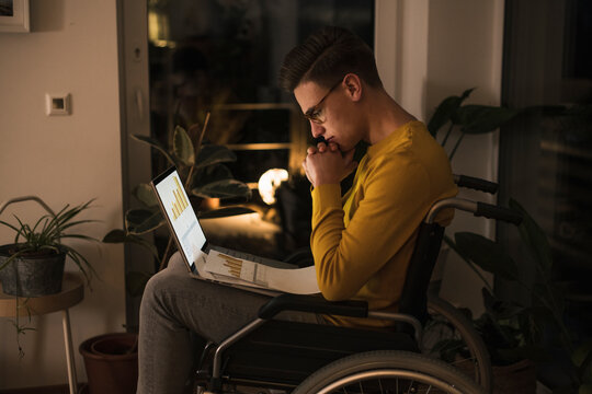 Concentrated oung man in wheelchair with laptop working late while sitting on wheelchair in living room