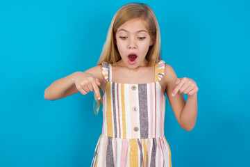 Amazed beautiful Caucasian little girl wearing stripped dress over blue background points down with...